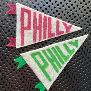 May include: Two felt pennants with the word "PHILLY" in bold letters. One pennant is white with red lettering and a red accent. The other is white with green lettering and a green accent. The pennants are triangular.