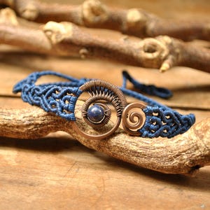 Peut inclure: Un bracelet macramé bleu fait à la main avec un motif en spirale en cuivre et une petite pierre précieuse bleue ronde. Le bracelet est présenté sur une branche en bois sur un fond en bois rustique.