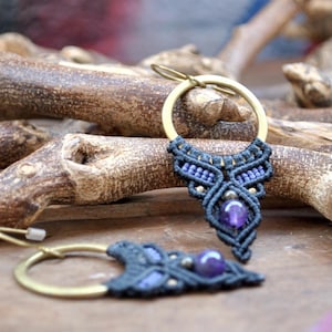 May include: A pair of handmade earrings with a dark blue macrame design and amethyst stones. The earrings are hanging from brass hoops.