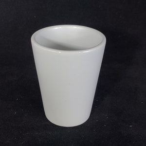 Puede incluir: Una taza de cerámica blanca con un borde liso y redondeado. La taza está vacía y se encuentra sobre una superficie negra.