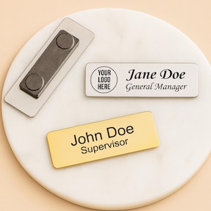 Puede incluir: Un conjunto de placas de identificación con cierres magnéticos. Una placa es dorada con "John Doe, Supervisor" impreso. Otra es blanca con "Jane Doe, General Manager" y un espacio para un logotipo. El cierre magnético es plateado.