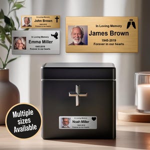 May include: A black cremation urn with a silver cross and a nameplate that reads "Noah Miller 1945-2019 Forever in our hearts." Additional nameplates with photos and text are displayed. A candle and vase are also present.