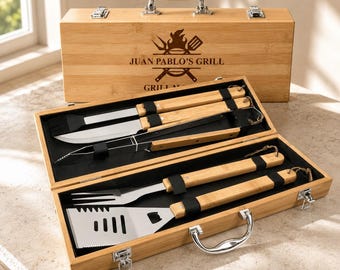 Puede incluir: Un juego de herramientas para parrilla de bambú en un maletín de madera rectangular con asa y cierres metálicos. La tapa presenta el texto grabado JUAN PABLO'S GRILL con un icono de llama y utensilios. Incluye herramientas de acero.