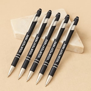 May include: Five black pens with silver accents and the text "YOUR TEXT HERE" printed on the barrel. The pens are arranged on a light-colored, rectangular stone. The pens have a sleek, modern design.