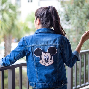 May include: A blue denim jacket with a Mickey Mouse patch on the back. The patch is black and white with a red nose and yellow eyes.