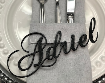 May include: A black wooden place card with the name "Adriel" written in cursive script. The place card is sitting on a gray linen napkin with a white lace trim. The napkin is folded and placed on a white plate with a silver rim.