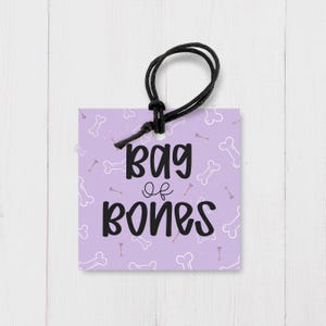Peut inclure: Une étiquette cadeau carrée lavande avec une boucle de cordon noire. L'étiquette présente un motif répété d'os de chien blancs et bruns. Le texte noir en gras indique "Bag of Bones".
