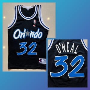 May include: Black basketball jersey with white and blue accents. The jersey features the word "Orlando" in white with a star graphic, and the number "32" in blue. The back of the jersey displays "O'NEAL" and the number "32".