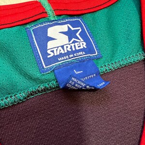 May include: Close-up of a vintage Starter athletic jersey. The jersey features a green, purple, and red color scheme. The Starter logo is prominently displayed on a blue tag, with the text "Made in Korea". A smaller tag indicates the size is Large.