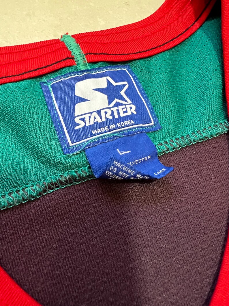 May include: Close-up of a vintage Starter athletic jersey. The jersey features a green, purple, and red color scheme. The Starter logo is prominently displayed on a blue tag, with the text "Made in Korea". A smaller tag indicates the size is Large.