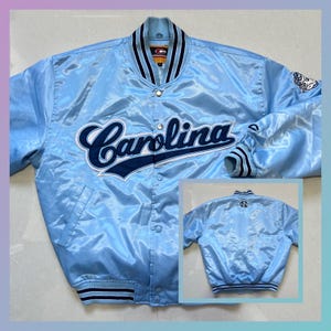 May include: Light blue satin jacket with the word "Carolina" in navy blue script across the chest. The jacket has a button-up front, navy blue and white striped cuffs and collar, and a small patch on the sleeve.