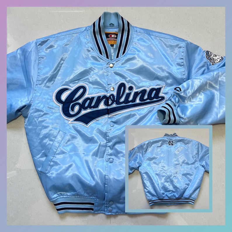 K&ouml;nnte beinhalten: Hellblaue Satinjacke mit dem Wort "Carolina" in marineblauer Schrift &uuml;ber der Brust. Die Jacke hat eine Knopfleiste, marineblau-wei&szlig; gestreifte B&uuml;ndchen und Kragen sowie einen kleinen Aufn&auml;her am &Auml;rmel.