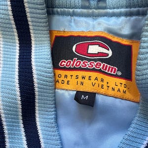 K&ouml;nnte beinhalten: Nahaufnahme einer hellblauen Jacke mit marineblauen und wei&szlig; gestreiften Zierleisten. Die Jacke hat ein Colosseum Sports wear-Etikett mit der Gr&ouml;&szlig;e M. Hergestellt in Vietnam.