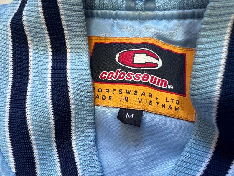 K&ouml;nnte beinhalten: Nahaufnahme einer hellblauen Jacke mit marineblauen und wei&szlig; gestreiften Zierleisten. Die Jacke hat ein Colosseum Sports wear-Etikett mit der Gr&ouml;&szlig;e M. Hergestellt in Vietnam.