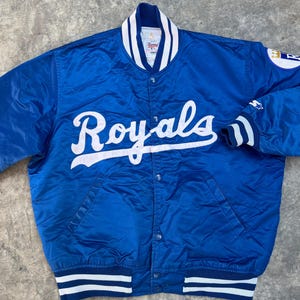 May include: A blue satin baseball jacket with white and blue striped ribbing. The jacket has a snap closure and features the word "Royals" in white script on the front.