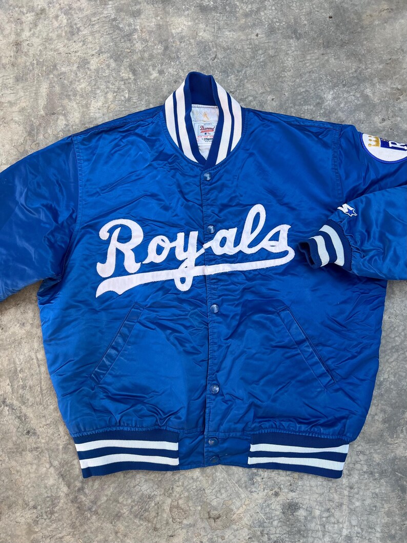May include: A blue satin baseball jacket with white and blue striped ribbing. The jacket has a snap closure and features the word "Royals" in white script on the front.