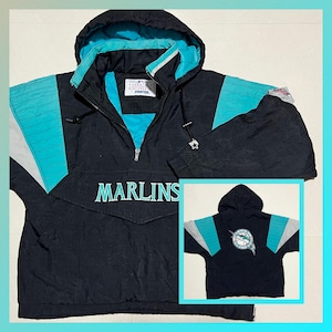 May include: Black and teal anorak jacket with the word "MARLINS" across the front. The jacket has a hood and a zipper closure. The sleeves have teal and gray accents. A smaller image of the jacket's back is in the lower right corner.