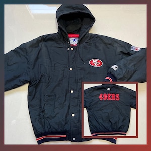Könnte beinhalten: Schwarze Kapuzenjacke mit dem Logo der San Francisco 49ers auf Brust und Ärmel. Die Jacke hat ein rot-weißes Logo und Zierleisten. Ein kleineres Bild zeigt die Rückseite einer schwarzen Jacke mit "49ERS" in Rot.