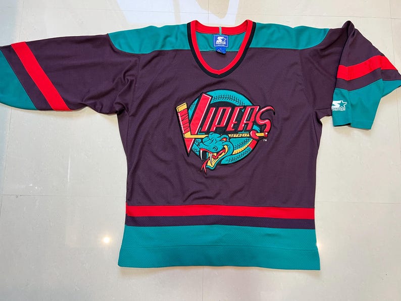 May include: A purple hockey jersey with teal and red accents. The jersey features the word "Vipers" in red and yellow, along with a teal snake logo. The jersey has a V-neck and is made by Starter.