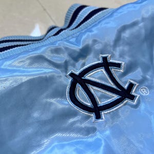 K&ouml;nnte beinhalten: Hellblaue Satinjacke mit marineblau-wei&szlig; gestreiftem Kragen. Mit einem marineblauen gestickten Logo mit den Buchstaben "NC" und einem Warenzeichen. Die Jacke hat eine gl&auml;nzende, glatte Textur.
