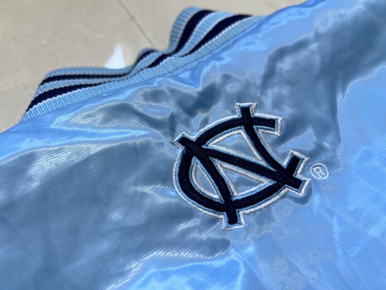 K&ouml;nnte beinhalten: Hellblaue Satinjacke mit marineblau-wei&szlig; gestreiftem Kragen. Mit einem marineblauen gestickten Logo mit den Buchstaben "NC" und einem Warenzeichen. Die Jacke hat eine gl&auml;nzende, glatte Textur.