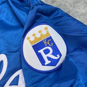 May include: A blue satin baseball jacket with a white embroidered letter "R" and a white patch with a gold crown and the letters "KC" on the right sleeve.