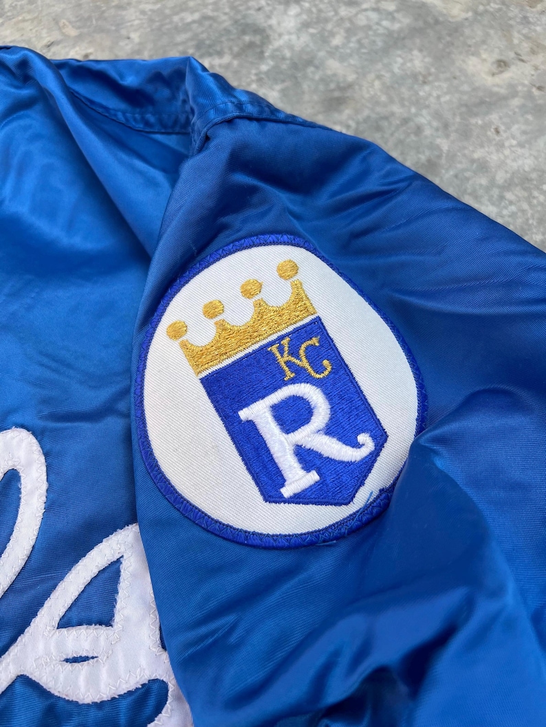 May include: A blue satin baseball jacket with a white embroidered letter "R" and a white patch with a gold crown and the letters "KC" on the right sleeve.