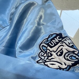 K&ouml;nnte beinhalten: Hellblaue Satinjacke mit einem Aufn&auml;her mit den Buchstaben "UNC" und einem Cartoon-Widder-Logo. Die Jacke hat eine gl&auml;nzende, glatte Textur. Der Aufn&auml;her ist wei&szlig;, marineblau und hellblau. Die Jacke ist wahrscheinlich Teil der Kleidung eines Sportteams.