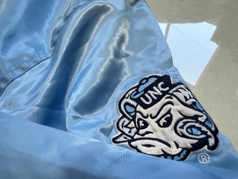 K&ouml;nnte beinhalten: Hellblaue Satinjacke mit einem Aufn&auml;her mit den Buchstaben "UNC" und einem Cartoon-Widder-Logo. Die Jacke hat eine gl&auml;nzende, glatte Textur. Der Aufn&auml;her ist wei&szlig;, marineblau und hellblau. Die Jacke ist wahrscheinlich Teil der Kleidung eines Sportteams.