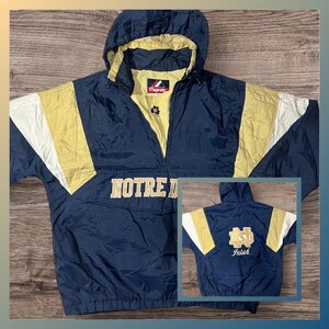 Puede incluir: Una chaqueta con capucha azul marino y dorada que presenta el texto NOTRE DAME en la parte delantera y un logotipo de Notre Dame con la palabra Irish en la parte posterior. La chaqueta tiene una etiqueta de marca Majestic en el cuello.