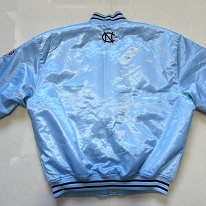 K&ouml;nnte beinhalten: Hellblaue Satinjacke mit schwarz-wei&szlig; gestreiftem Kragen und Manschetten. Auf der R&uuml;ckseite der Jacke befindet sich ein schwarz-wei&szlig;es Logo. Die Jacke hat einen Druckknopfverschluss und lange &Auml;rmel.