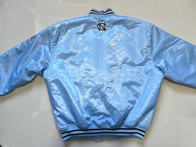 K&ouml;nnte beinhalten: Hellblaue Satinjacke mit schwarz-wei&szlig; gestreiftem Kragen und Manschetten. Auf der R&uuml;ckseite der Jacke befindet sich ein schwarz-wei&szlig;es Logo. Die Jacke hat einen Druckknopfverschluss und lange &Auml;rmel.