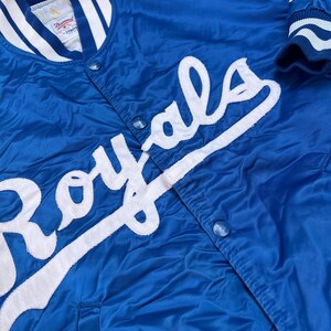 May include: A blue satin baseball jacket with white stitching and the word "Royals" in white lettering on the front.