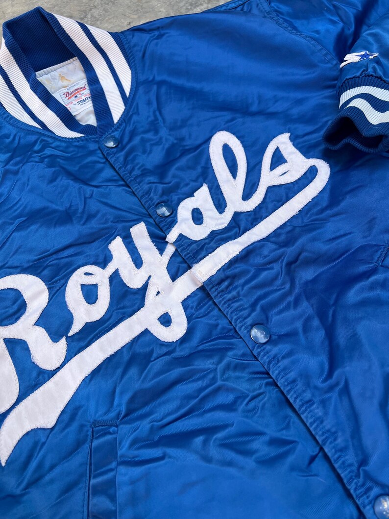 May include: A blue satin baseball jacket with white stitching and the word "Royals" in white lettering on the front.