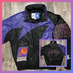 May include: A black and purple leather jacket with the Phoenix Suns logo on the front and back. The jacket has a zipper closure and a stand-up collar.