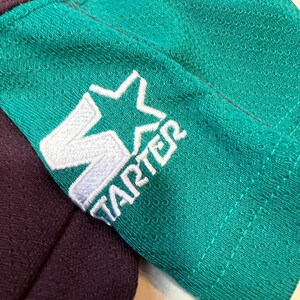 May include: Close-up of a teal and purple athletic jersey sleeve. The teal sleeve features a white embroidered Starter logo. The jersey has a mesh texture. The purple fabric is visible in the background.