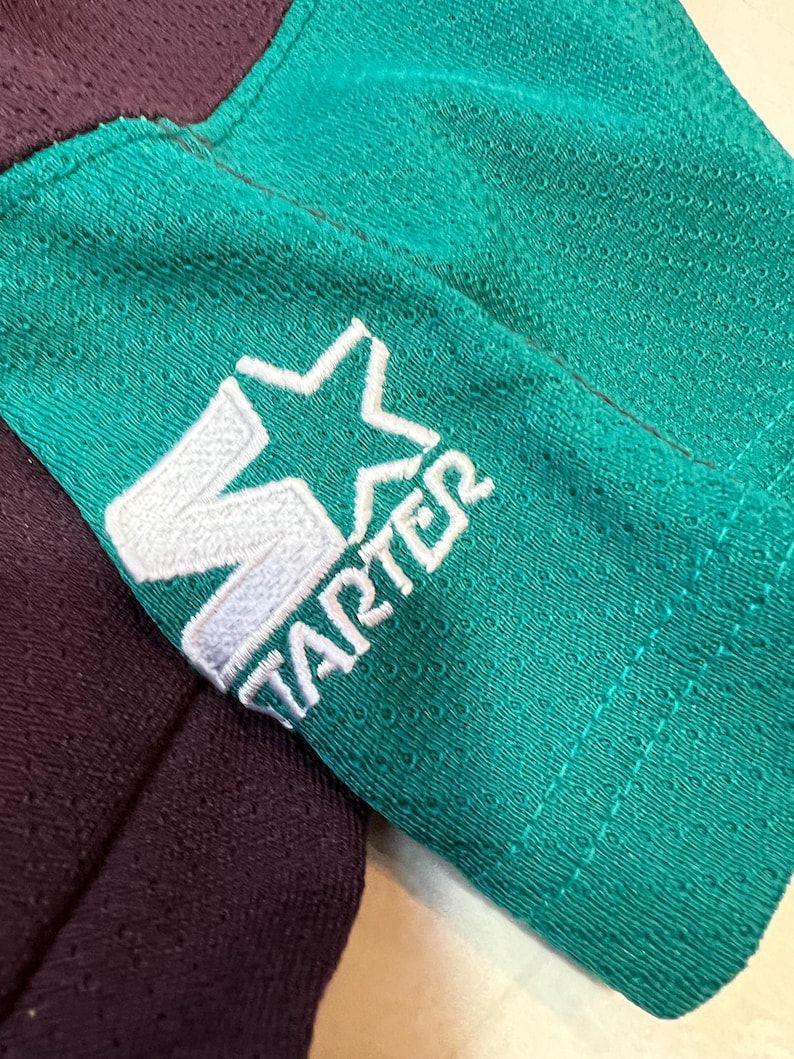 May include: Close-up of a teal and purple athletic jersey sleeve. The teal sleeve features a white embroidered Starter logo. The jersey has a mesh texture. The purple fabric is visible in the background.