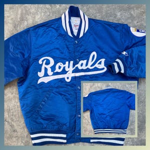 May include: A blue satin baseball jacket with white lettering that says "Royals" on the front. The jacket has a white and blue striped collar and cuffs. The back of the jacket is also blue with a white and blue striped waistband.