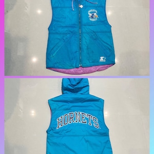 Puede incluir: Un chaleco reversible turquesa y morado con capucha. La parte delantera presenta el texto "CHARLOTTE HORNETS" y un logotipo. La parte trasera del chaleco muestra la palabra "HORNETS" en letras grandes. El chaleco tiene cierre de cremallera.