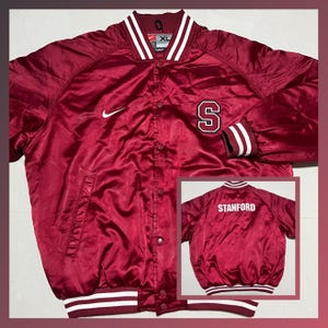 May include: Maroon satin bomber jacket with white and maroon striped cuffs and collar. Features a white Nike swoosh and a large "S" patch. The jacket has a snap-button closure and a small inset image of the jacket's back with the word "STANFORD".