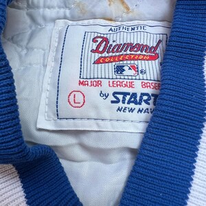 May include: A white baseball jacket with blue trim and a white patch that says "Authentic Diamond Collection Major League Baseball by Start New York".