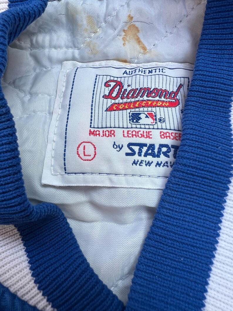 May include: A white baseball jacket with blue trim and a white patch that says "Authentic Diamond Collection Major League Baseball by Start New York".