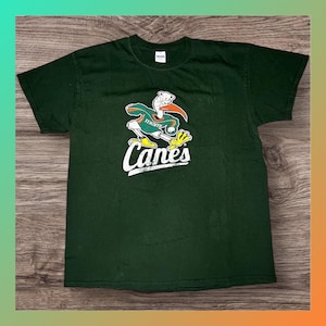 Pode incluir: T-shirt verde escuro com um mascote de pássaro de desenho animado com detalhes em laranja, verde e branco. A palavra "Miami" está acima do pássaro, e a palavra "Canes" está escrita em branco abaixo.