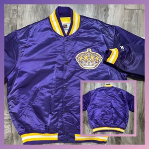 Könnte beinhalten: Eine lilafarbene Satin-Baseballjacke mit einem gelb-weiß gestreiften Kragen und Bündchen. Die Jacke hat ein großes gesticktes Wappen auf der linken Brust und ein kleines gesticktes "M" auf dem rechten Ärmel.