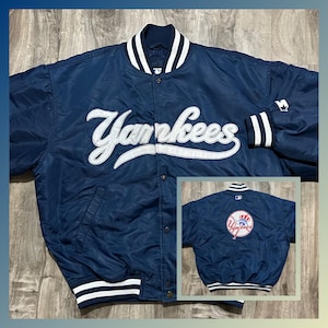 Puede incluir: Una chaqueta de béisbol de satén azul marino con rayas blancas en las mangas y el cuello. La chaqueta tiene un gran logotipo "Yankees" bordado en blanco en la parte delantera y un logotipo bordado más pequeño en la parte posterior.