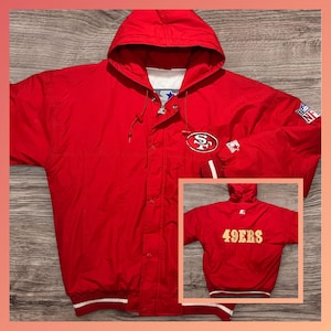 Könnte beinhalten: Rote Kapuzenjacke mit dem Logo der San Francisco 49ers auf Brust und Ärmel. Die Jacke hat einen weiß-rot gestreiften Saum und Bündchen. Auf der Rückseite der Jacke steht der Schriftzug "49ERS" in Gold.