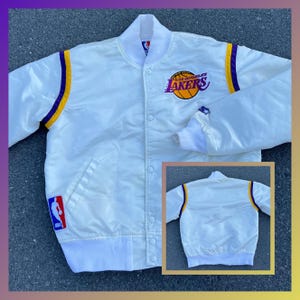 Könnte beinhalten: Weiße Satin-Bomberjacke mit violetten und goldenen Akzenten. Die Jacke trägt das Logo der Los Angeles Lakers auf der Vorderseite und das NBA-Logo auf dem linken Ärmel.