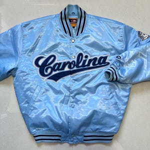 K&ouml;nnte beinhalten: Hellblaue Satinjacke mit dem Wort "Carolina" in marineblauer Schrift auf der Brust. Die Jacke hat eine Knopfleiste, gestreifte Manschetten und einen Kragen mit marineblauen Streifen. Ein Logo ist auf dem rechten &Auml;rmel sichtbar.