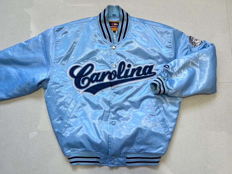 K&ouml;nnte beinhalten: Hellblaue Satinjacke mit dem Wort "Carolina" in marineblauer Schrift auf der Brust. Die Jacke hat eine Knopfleiste, gestreifte Manschetten und einen Kragen mit marineblauen Streifen. Ein Logo ist auf dem rechten &Auml;rmel sichtbar.