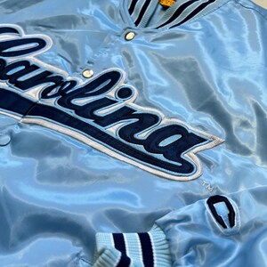 K&ouml;nnte beinhalten: Hellblaue Satinjacke mit dem Wort "Carolina" in Marineblau und Wei&szlig; gestickt. Die Jacke hat einen gerippten Kragen und Manschetten mit marineblauen und wei&szlig;en Streifen. Die Jacke hat einen Druckknopfverschluss.